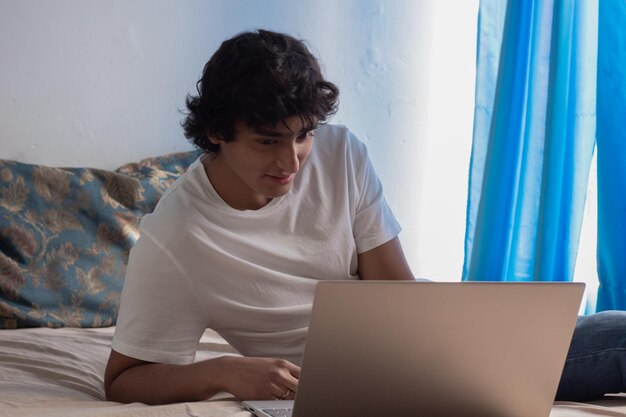 Sorrindo jovem com cabelo encaracolado, deitado na cama, trabalhando em seu laptop em casa