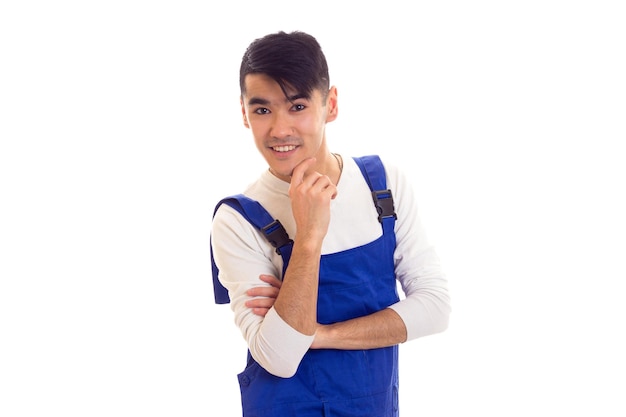 Sorrindo jovem com cabelo escuro, vestindo camisa branca e macacão azul sobre fundo branco no estúdio