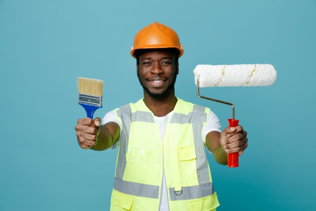 Sorrindo jovem construtor americano africano de uniforme segurando pincel de rolo com pincel para câmera isolada em fundo azul