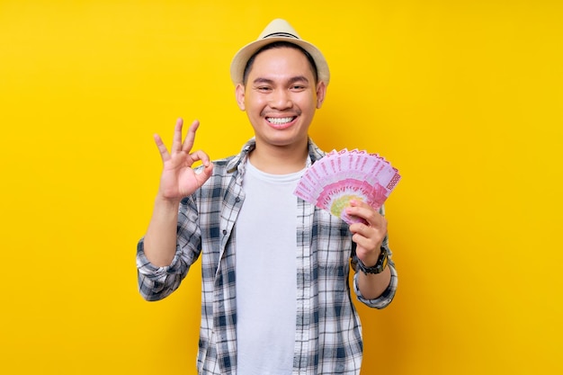 Sorrindo jovem de etnia asiática 20 anos vestindo roupas casuais chapéu segurando na mão leque de dinheiro em notas de rupiah mostra ok ok gesto isolado no fundo amarelo Pessoas conceito de estilo de vida