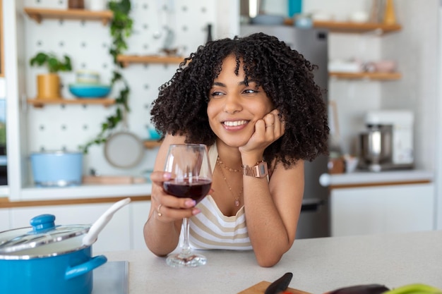 Sorrindo jovem dona de casa com copo de vinho tinto enquanto ela está no fogão na cozinha preparando o jantar