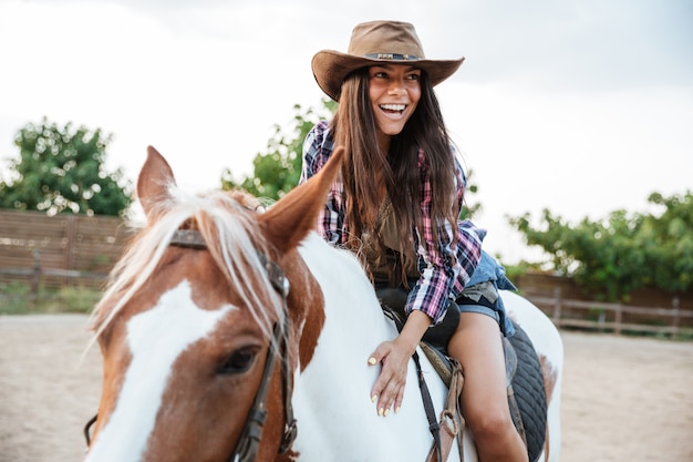 Foto sorrindo, jovem e fofa cowgirl womna andando a cavalo ao ar livre e rindo