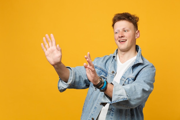 Sorrindo jovem em roupas casuais jeans, olhando de lado em pé com espalhando as mãos estendidas isoladas em fundo amarelo alaranjado. conceito de estilo de vida de emoções sinceras de pessoas. mock up espaço de cópia.