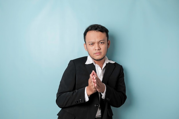Sorrindo jovem empresário asiático vestindo terno preto gesticulando saudação ou namaste isolado sobre fundo azul