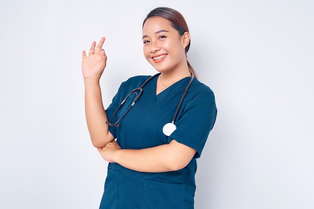 Sorrindo jovem enfermeira asiática vestindo uniforme azul com estetoscópio garante a qualidade dos resultados dos testes em seu laboratório clínico mostrando sinal ok isolado no fundo branco