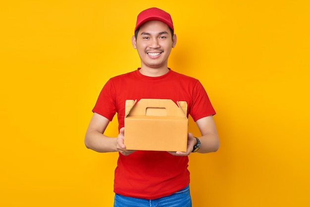 Sorrindo jovem entregador asiático em roupa de trabalho de camiseta de boné vermelho trabalha como correio de revendedor segurando sobremesa de bolo em caixa vazia sem marcação isolada em fundo amarelo Conceito de serviço