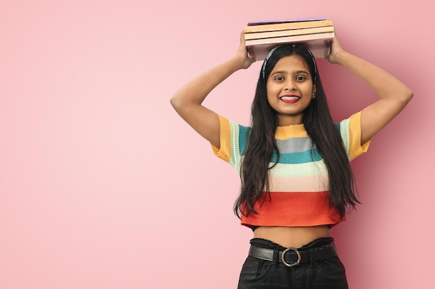 Sorrindo jovem estudante asiática indiana posando livros de equilíbrio isolados na cabeça olhando diretamente para a câmera mulher de cabelos escuros expressando emoções positivas