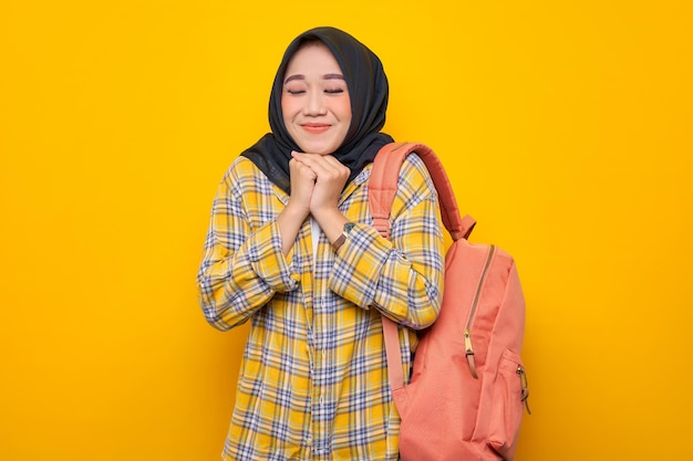 Sorrindo jovem estudante muçulmana asiática em roupas casuais e mochila fazendo um gesto de oração com os olhos fechados isolados em fundo amarelo conceito de faculdade universitária de escola de educação