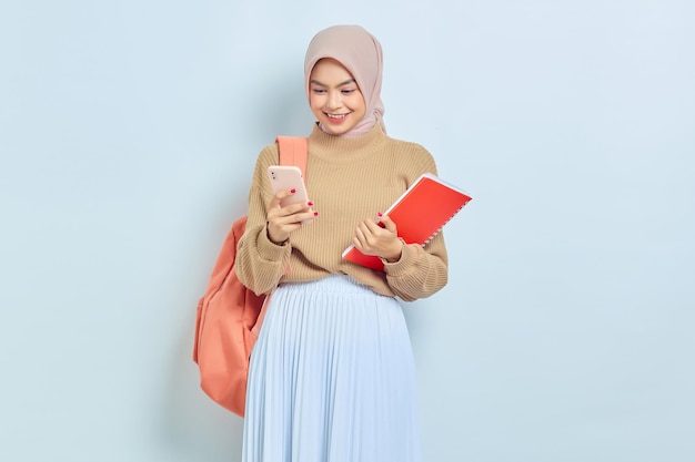 Sorrindo jovem estudante muçulmana asiática em suéter marrom com mochila segurando livro e telefone celular isolado em fundo branco de volta ao conceito de escola