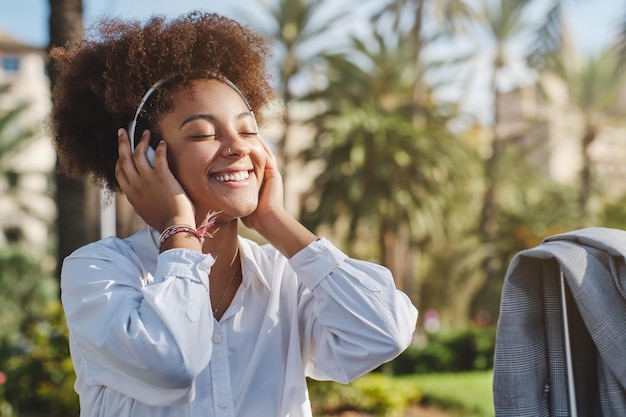 Sorrindo jovem étnica em fones de ouvido curtindo músicas em dia de verão brilhante no parque da cidade verde com palmeiras no fundo desfocado