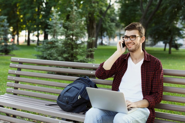 Sorrindo jovem falando no celular, trabalhando no laptop, sentado no parque. Tecnologia, comunicação, educação e conceito de trabalho remoto, espaço de cópia