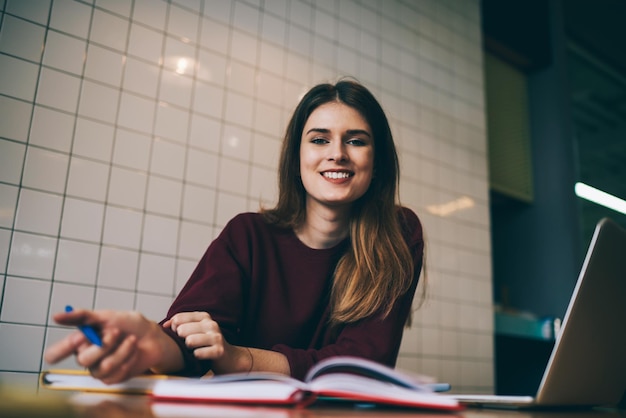 Sorrindo jovem freelancer profissional sentado perto do computador portátil e olhando para a câmera