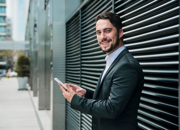 Foto sorrindo, jovem, homem negócios fica, exterior, a, edifício escritório, segurando móvel, em, mão