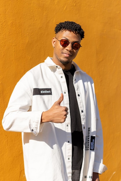 sorrindo jovem latino com óculos de sol posando olhando para a câmera e fazendo o símbolo de eu gosto com a mão, fundo amarelo vertical