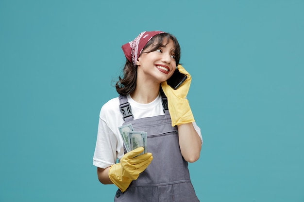 Sorrindo jovem limpador feminino vestindo bandana uniforme e luvas de borracha segurando dinheiro olhando para o lado enquanto fala no telefone isolado em fundo azul