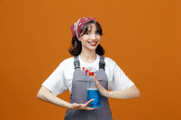 Sorrindo jovem limpador feminino vestindo uniforme e bandana segurando limpador com as duas mãos olhando para câmera isolada em fundo laranja