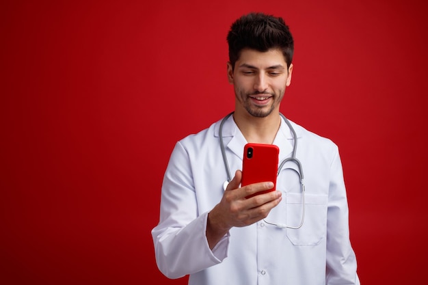 Sorrindo jovem médico masculino vestindo uniforme médico e estetoscópio em volta do pescoço segurando e olhando para o celular isolado em fundo vermelho com espaço de cópia