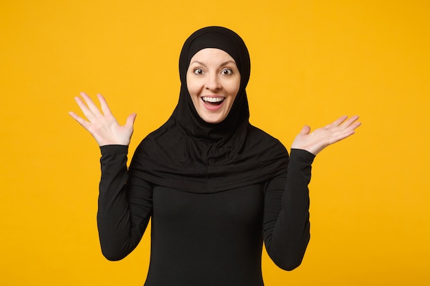 Sorrindo jovem muçulmana árabe em roupas pretas de hijab, espalhando as mãos, isoladas na parede amarela, retrato. Conceito de estilo de vida religioso de pessoas.