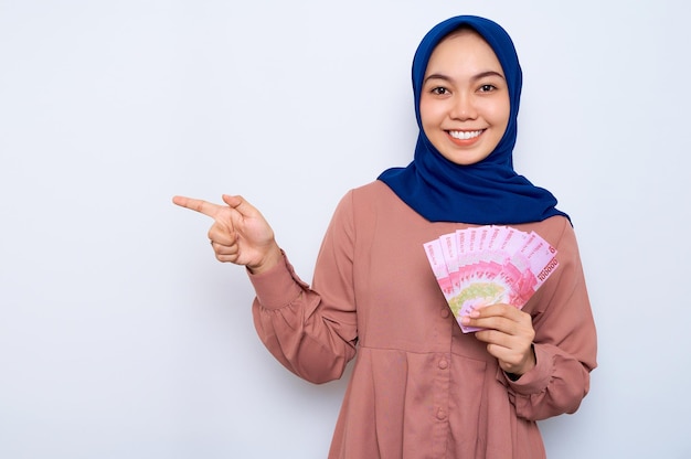 Sorrindo jovem muçulmana asiática na camisa rosa segurando notas de dinheiro e apontando os dedos para o espaço em branco isolado sobre fundo branco Pessoas conceito de estilo de vida religioso