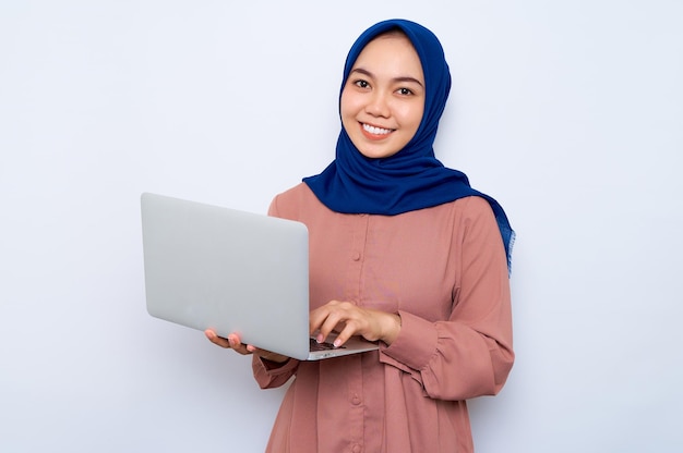 Sorrindo jovem muçulmana asiática na camisa rosa segurando um laptop e olhando para a câmera isolada sobre fundo branco Conceito de estilo de vida religioso de pessoas