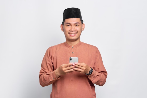 Sorrindo jovem muçulmano asiático usando um telefone celular e olhando para a câmera isolada sobre fundo branco