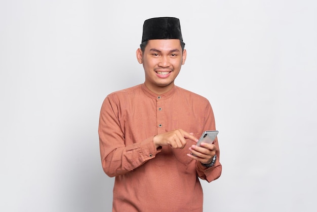 Sorrindo jovem muçulmano asiático usando um telefone celular e olhando para a câmera isolada sobre fundo branco