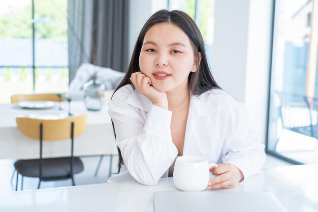 sorrindo jovem mulher asiática na camisa branca com uma xícara de chá ou café e laptop em casa moderna