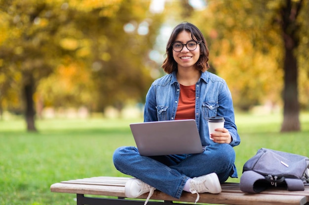 Sorrindo jovem mulher do Oriente Médio usando laptop e tomando café ao ar livre