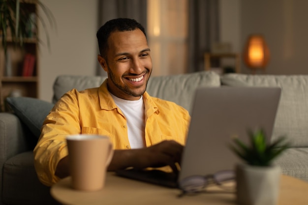 Foto sorrindo jovem negro trabalhando ou se comunicando online sentado no sofá com laptop em casa