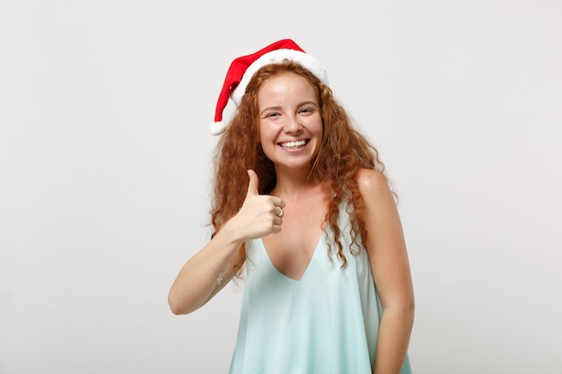 Sorrindo jovem ruiva santa em roupas leves, chapéu de natal, isolado no fundo branco, retrato de estúdio. feliz ano novo conceito de feriado de celebração de 2020. simule o espaço da cópia. mostrando o polegar para cima.