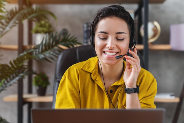 Sorrindo jovem teleoperadora trabalhando em home office com laptop e fone de ouvido