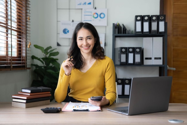 Sorrindo linda empresária asiática analisando gráfico e gráfico mostrando mudanças no mercado e segurando smartphone no escritório