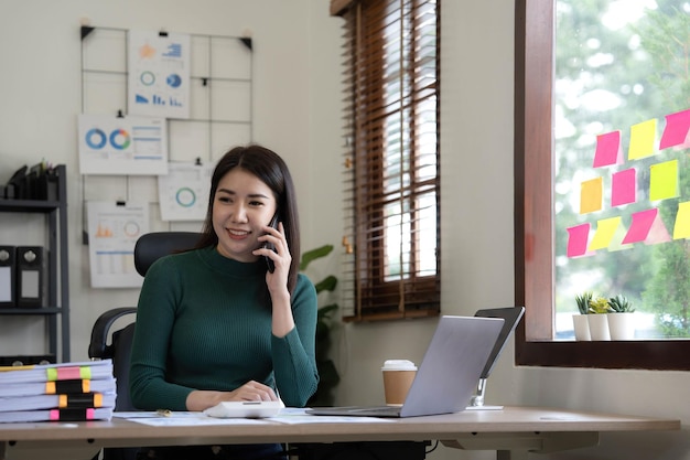 Sorrindo linda empresária asiática analisando gráfico e gráfico mostrando mudanças no mercado e segurando smartphone no escritório