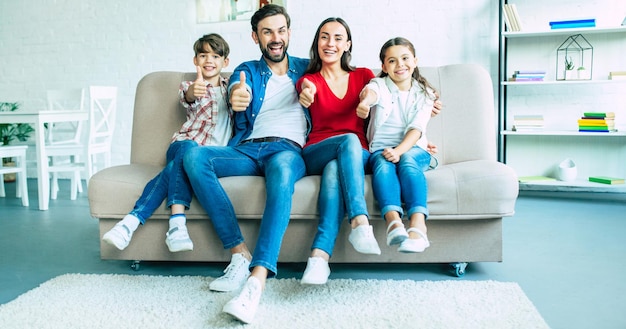 Sorrindo linda família moderna está feliz e mostra os polegares na câmera enquanto está sentado no sofá