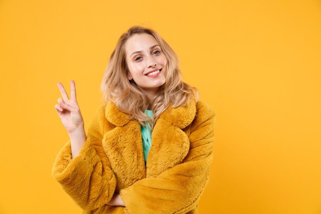 Sorrindo linda jovem loira com casaco de pele amarelo posando isolado no retrato de estúdio de fundo de parede laranja. Conceito de estilo de vida de emoções sinceras de pessoas. Mock up espaço de cópia. Mostrando sinal de vitória.