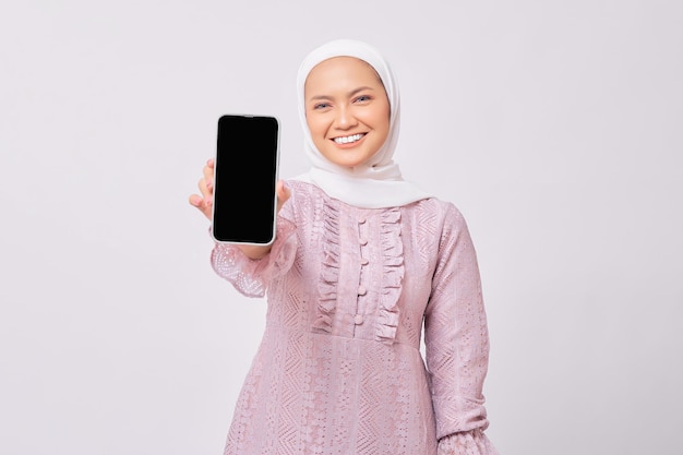 Sorrindo linda jovem muçulmana asiática usando hijab e vestido roxo, mostrando o telefone móvel com tela em branco, isolada no fundo branco do estúdio