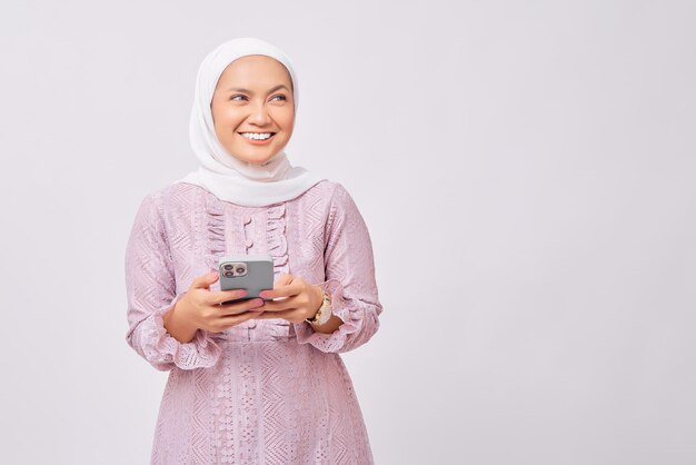 Sorrindo linda jovem muçulmana asiática usando hijab e vestido roxo segurando o telefone móvel e olhando para o espaço livre para propaganda isolada no fundo branco do estúdio