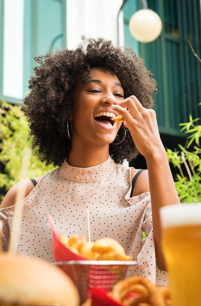 Foto sorrindo linda jovem negra feliz com piercing no nariz comendo fatias de batata em um pub em uma vista de ângulo baixo sobre comida e copos de cerveja