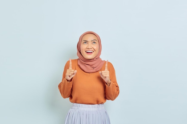 Sorrindo linda mulher asiática de suéter marrom e hijab apontando o dedo no espaço da cópia mostrando produtos publicitários e confiante olhando para a câmera isolada sobre fundo branco