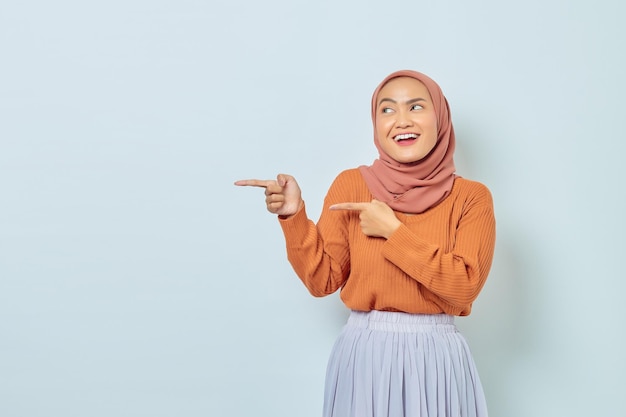 Sorrindo linda mulher asiática de suéter marrom e hijab apontando o dedo no espaço da cópia mostrando produtos publicitários e confiante olhando para a câmera isolada sobre fundo branco