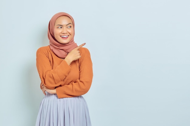 Sorrindo linda mulher asiática de suéter marrom e hijab apontando o dedo no espaço da cópia mostrando produtos publicitários e confiante olhando para a câmera isolada sobre fundo branco