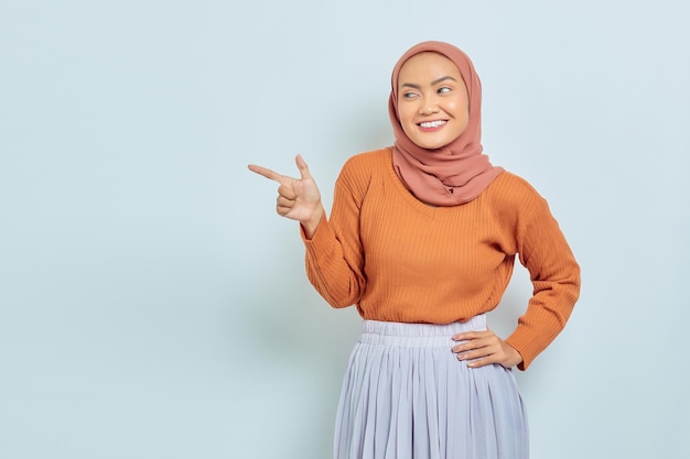Sorrindo linda mulher asiática de suéter marrom e hijab apontando o dedo no espaço da cópia mostrando produtos publicitários e confiante olhando para a câmera isolada sobre fundo branco