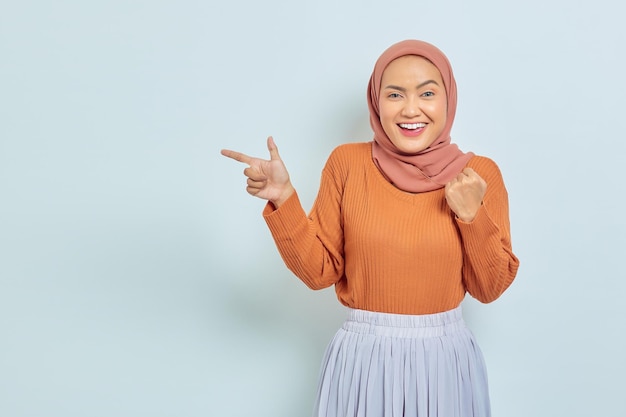 Sorrindo linda mulher asiática de suéter marrom e hijab apontando o dedo no espaço da cópia mostrando produtos publicitários e confiante olhando para a câmera isolada sobre fundo branco