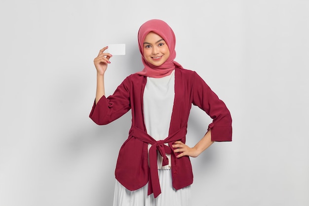 Sorrindo linda mulher asiática em camisa casual e hijab mostrando cartão de crédito, mão na cintura isolada sobre fundo branco. Conceito de estilo de vida religioso de pessoas