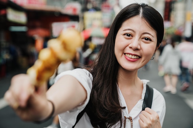 Sorrindo linda mulher coreana asiática segurando espetos de ovos mostrando para a câmera com rosto encantador. feliz adorável fêmea em pé ao ar livre tentando comida de rua local tradicional no mercado em dia ensolarado