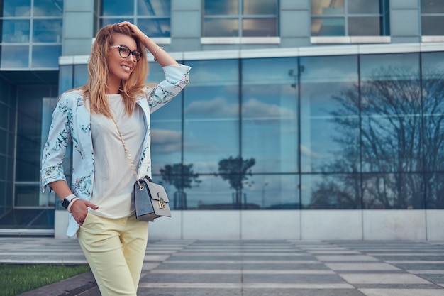 Sorrindo linda mulher elegante em roupas elegantes e óculos com uma bolsa, de pé contra um arranha-céu.