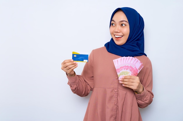 Sorrindo linda mulher muçulmana asiática em uma camisa casual e hijab segurando notas de rúpia indonésia mostrando cartão de crédito isolado sobre fundo branco Conceito de estilo de vida religioso de pessoas