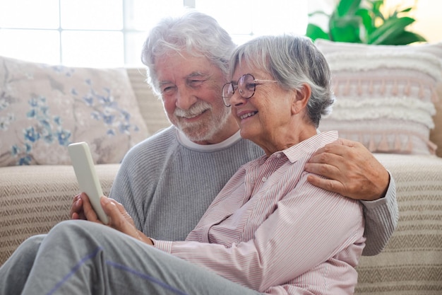 Sorrindo lindo casal sênior caucasiano sentado no chão em casa usando telefone inteligente velhos e conceito de novas tecnologias