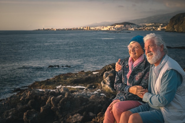 Sorrindo lindo casal sênior sentado abraçando nas rochas no mar desfrutando da luz do pôr do sol Estilo de vida descontraído para um casal caucasiano em férias ou aposentadoria