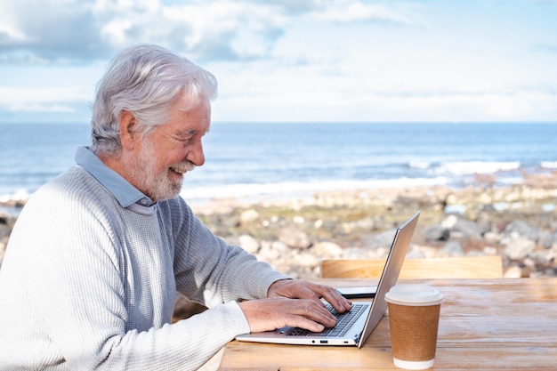 Sorrindo lindo homem sênior maduro digitando no teclado do computador ao ar livre sentado na mesa de madeira